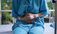 Patient in a blue hospital gown sitting on a bed, holding their stomach, indicating discomfort or pain. Relevant to healthcare discussions.